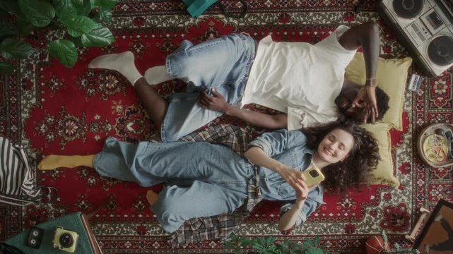 Young Black man and Caucasian woman making funny faces and taking selfies with phone then laughing while relaxing on red retro carpet surrounded 90s vintage items. Directly above, zoom in shot