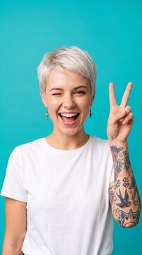 Cheerful young woman with short blonde hair and arm tattoos smiling and winking while showing a peace sign gesture against a bright blue background