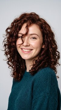 Radiant young woman with curly auburn hair and a warm friendly smile captured in a professional studio portrait against a clean light grey backdrop