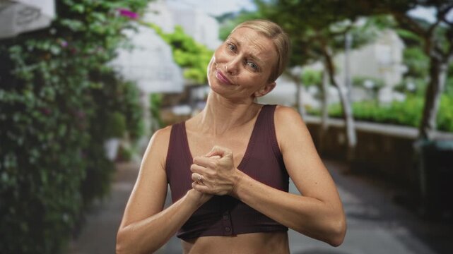 Woman middle aged blonde clasping hands close to chest, bare shoulders visible, wearing a maroon sportsbra on a street lined with trees and planters; gentle contentment.