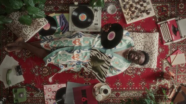 High angle shot of smiling Black man in patterned robe lying on red vintage rug, holding vinyl records and enjoying music next to retro turntable and chessboard. Zoom in