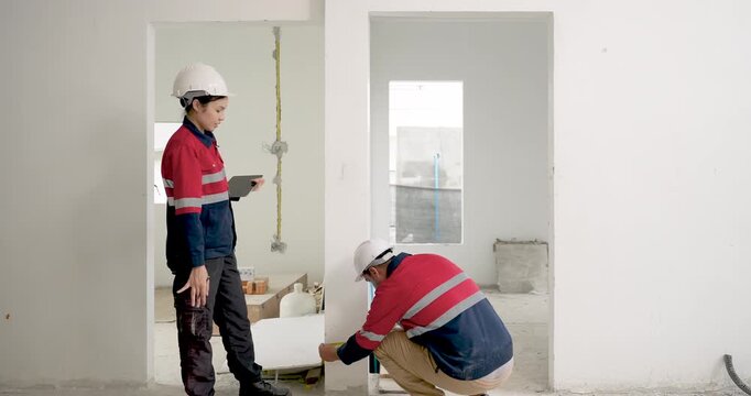 Asian female engineer observes male worker crouching near wall base, inspecting construction quality issue, safety compliance, renovation progress, site document checking, supervisor workflow indoors.