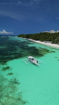 Vertical drone tracks a longboat moving along White Beach, showing turquoise shallows, coral reef, and palm-lined shore in Panglao