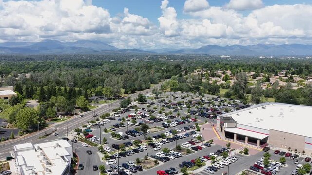 Packed parking lot surrounds large red-trimmed retail store in Redding, busy street stretches past scattered trees toward distant hazy mountains under partly cloudy sky. Aerial drone wide shot