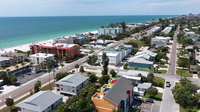 Rows of beachfront homes line Indian Rocks Beach street, turquoise Gulf waters meet white sand while palm-lined roads stretch toward distant coastal high-rise buildings. Aerial drone flyover shot