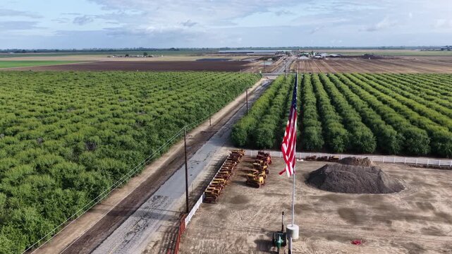 American flag flies proudly above farm equipment yard near Chowchilla, rows of green orchard trees stretch alongside dirt road toward distant Central Valley farmland. Aerial drone approaching shot