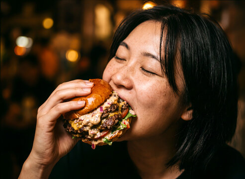 Close up of a happy Asian woman with freckles taking a big bite of a huge double cheeseburger with dripping sauce against a warm bokeh background.