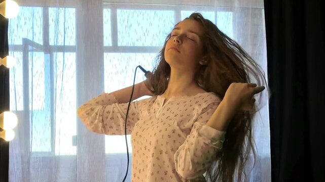 Teenage girl dries her long hair at home near a window, daily beauty routine, hair care, personal grooming, casual lifestyle.