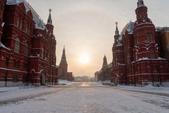 Snow-covered red brick buildings with spires and towers lining a wide avenue, illuminated by a low winter sun.