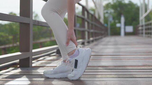 Runner woman with ankle injury pain rests during fitness running, seeking physical relief on outdoor wooden bridge to recover from exercise muscle strain on injured leg.