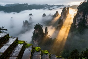 Fototapeta premium Mist-covered mountain peaks with sunbeams piercing through clouds, viewed from a mossy stone pathway