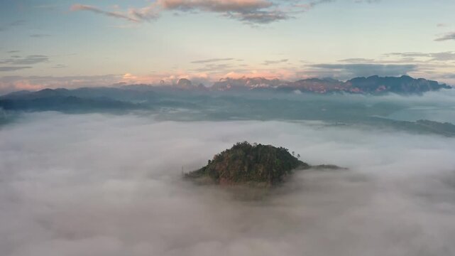 Tropical forest island peak rising over the sea of morning mist during a colorful sunrise over the dense jungle and mountains of Khao Sok National Park in Thailand. Aerial view drone flight panorama
