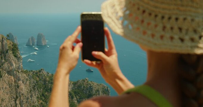 Woman in hat photographs Capri seascape with boats and distant Faraglioni sea stacks under clear sky