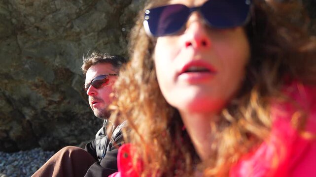 Couple conversation beach featuring a man and woman in sunglasses talking while sitting on a rocky shore during a sunny day