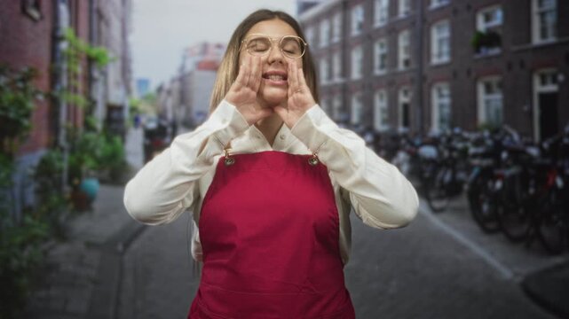 Woman in red apron cupping hands to shout, hands framing mouth on a city street lined with bicycles and brick buildings; community outreach determination.