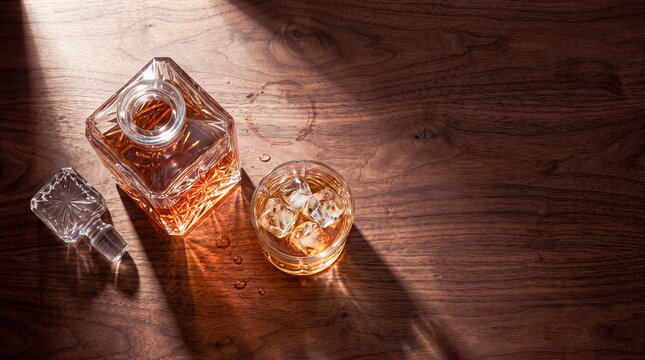 A Toast to Solitude: A crystal decanter of amber liquid and a chilled glass filled with ice and the same beverage sit on a polished wooden surface, bathed in dramatic sunlight and shadow.
