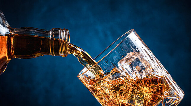 Amber Elixir Poured: Rich amber liquid, likely whiskey, is poured from a clear bottle into a heavy glass filled with ice cubes, set against a dark, moody backdrop.