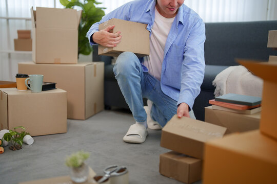 Asian man unpacking cardboard box while organizing belongings during home relocation in bright living room