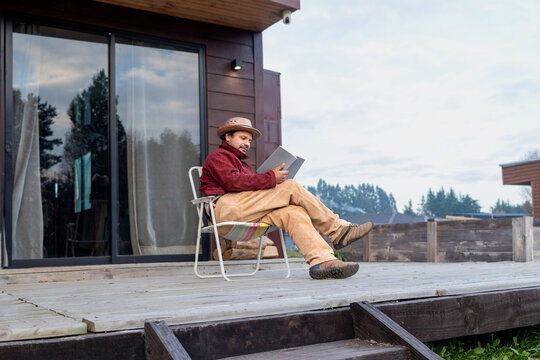 Man in a straw hat reading a book while relaxing on a wooden deck outside a modern cabin