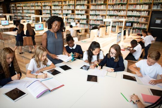 Diverse group of students and a teacher in a library, working on tablets.