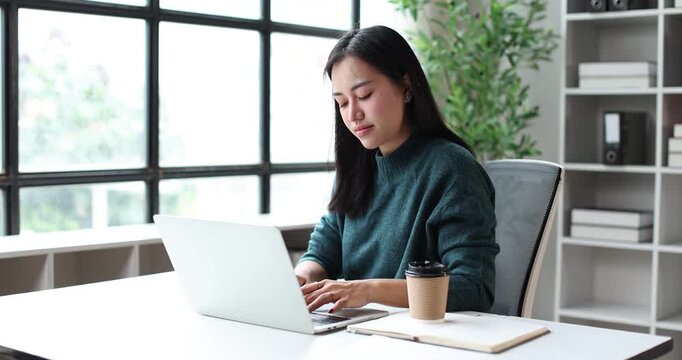 Asian woman feeling excitement and celebrating achievement while reading good news on a laptop computer, working remotely from her modern home office space
