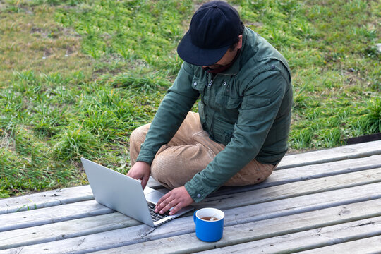 Man using a portable computer while sitting cross-legged outdoors on a cottage terrace
