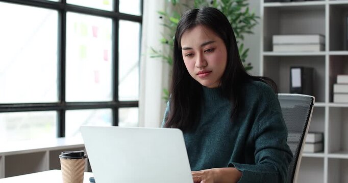 Asian woman feeling excitement and celebrating achievement while reading good news on a laptop computer, working remotely from her modern home office space