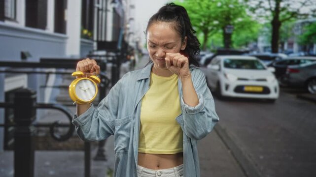 Woman holding yellow alarm clock and rubbing eye on street in urban morning wakeup scene; morning routine tired.