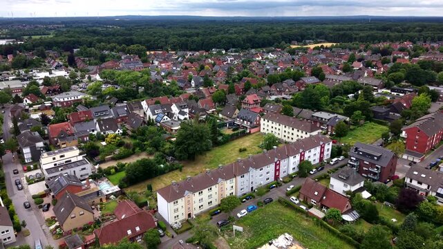 White multi-story apartment row stretches through residential German town, red-roofed houses cluster among gardens beside quiet street lined with parked cars plus trees. Aerial drone flyover shot