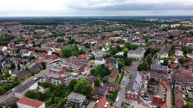 Dense red-roofed houses spread across historic town center in Dulmen, Germany, brick church spire rises above tree-lined cobblestone streets beneath overcast summer sky. Aerial drone flyover shot