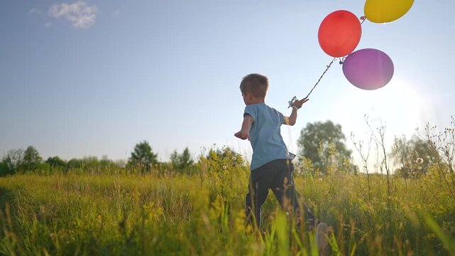 Boy runs with colorful balloons in meadow field. Child playing outdoor with balloon at sunset. Kid running in nature with balloons. Happy childhood in countryside with child and balloon play.