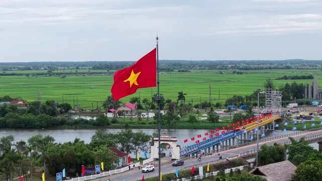 Wide aerial drone shot showing the Vietnamese flag flying high near the colorfully decorated Hien Luong Bridge, with the Ben Hai River and green countryside beyond.