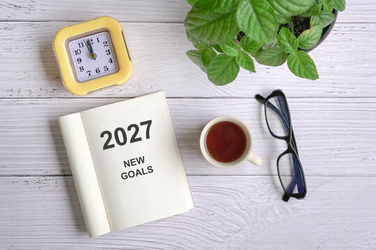 2027 New Goals, message on notepad next to coffee, eyeglasses, clock and green plant. Top view.