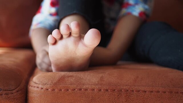 A little child struggles with foot pain on sofa with child's hand gently touching foot and showing discomfort in a close up motion clip