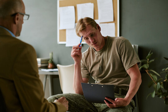 Young Caucasian man in military hospital reviewing clipboard during counseling session, reflecting on treatment and recovery. Useful for veteran healthcare, trauma support, rehabilitation marketing
