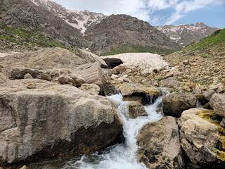 A rocky mountain waterfall cascades over moss-covered boulders near a snow cave, with patches of avalanche snow and rugged snow-capped peaks rising behind green hillsides. 📍 Hawraman Takht, Kurdistan © Hawraman83