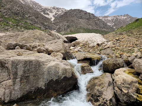 A rocky mountain waterfall cascades over moss-covered boulders near a snow cave, with patches of avalanche snow and rugged snow-capped peaks rising behind green hillsides.
📍 Hawraman Takht, Kurdistan