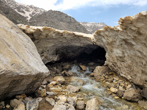 A large snow cave arches over a rocky mountain stream beside a huge boulder, opening onto snow-capped rugged peaks under a blue sky.
📍 Hawraman Takht, Kurdistan, Iran 