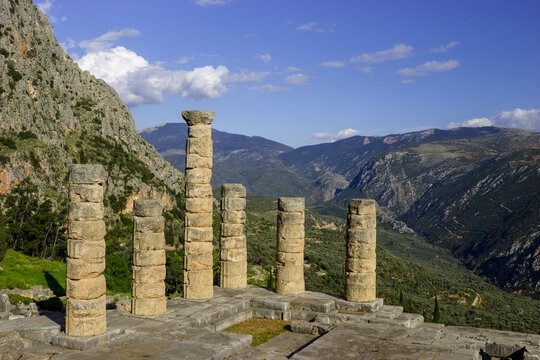 The Temple of Apollo at Delphi, Greece, with its ancient columns standing against a backdrop of mountains and a blue sky with clouds.