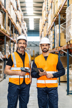 Two male warehouse workers wearing safety vests and hard hats, holding a barcode scanner and a digital tablet, standing in a large distribution center aisle