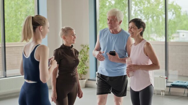 Active Caucasian seniors talking with young female fitness instructor in bright gym studio. Video is perfect for promoting healthy aging and senior wellness programs