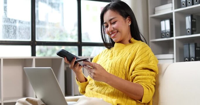 Woman sitting at home, holding a smartphone and credit card, engaged in online shopping and making secure mobile payments, utilizing digital banking for convenient purchases