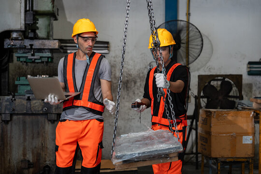 caucasian logistic manager using laptop to gesture instructions an asian colleague who is operating the chain hoist crane to lift wrapped pallet with cargo in industrial factory setting