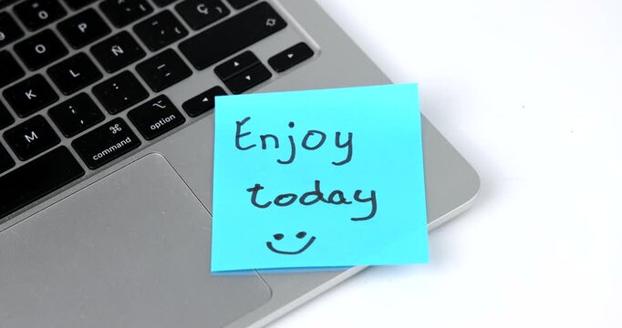 A handwritten blue sticky note saying "Enjoy today" is attached to a silver laptop. Horizontal composition representing positive thinking, organization and an inspiring office environment.