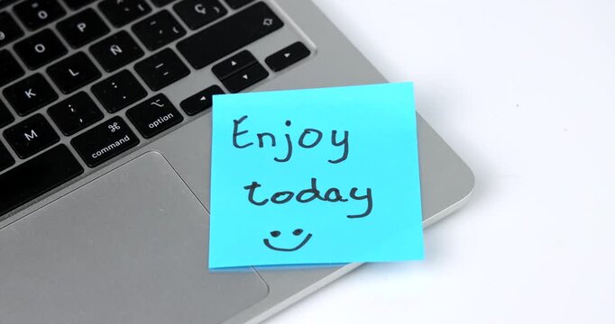 Blue adhesive note with the message "Enjoy today" placed on a silver laptop symbolizes encouragement, balance and productivity. Horizontal composition with clean minimalist office style.