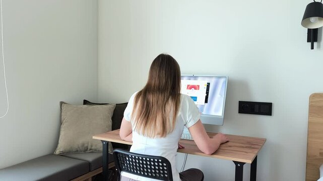 Woman sits at her home office desk, back view, navigating open windows on her computer display. She opens and closes software applications while working through tasks. A freelancer stays