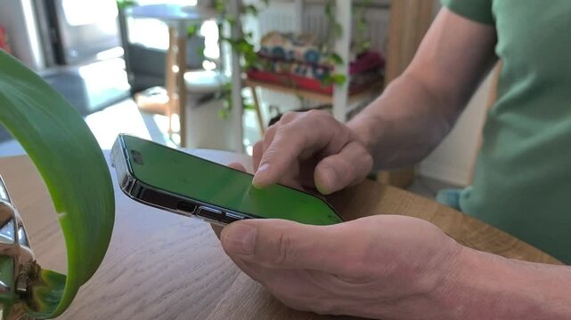 Man using smartphone at a wooden table indoors. Close up hands tap and scroll on mobile screen in a home interior. Casual digital browsing and communication..