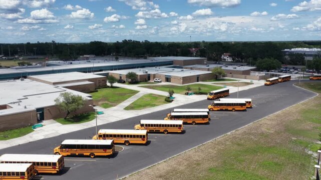 Yellow school buses queue in long rows across paved parking lot outside high school campus in Plant City, Florida, tan brick buildings sit beneath scattered white clouds. Aerial drone rising wide shot