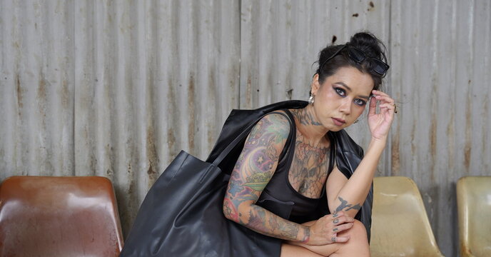 Young heavily tattooed Asian woman with sunglasses on her head sits holding a large black tote bag, looking at the camera with a confident expression