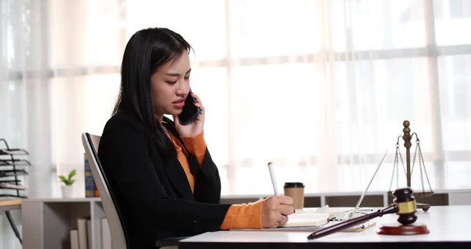 Young asian woman lawyer managing her legal work, talking on a mobile phone and writing important details in a notebook, symbolizing dedicated professional service and communication